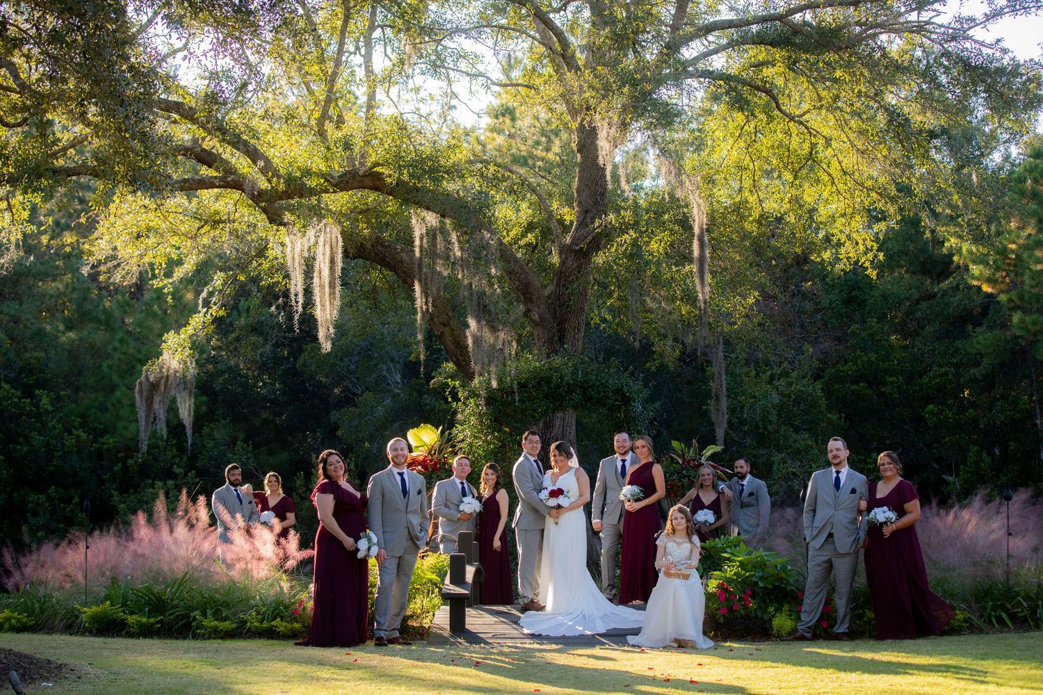 A wedding party poses together under moss-draped oak trees during golden hour in a scenic outdoor garden setting.
