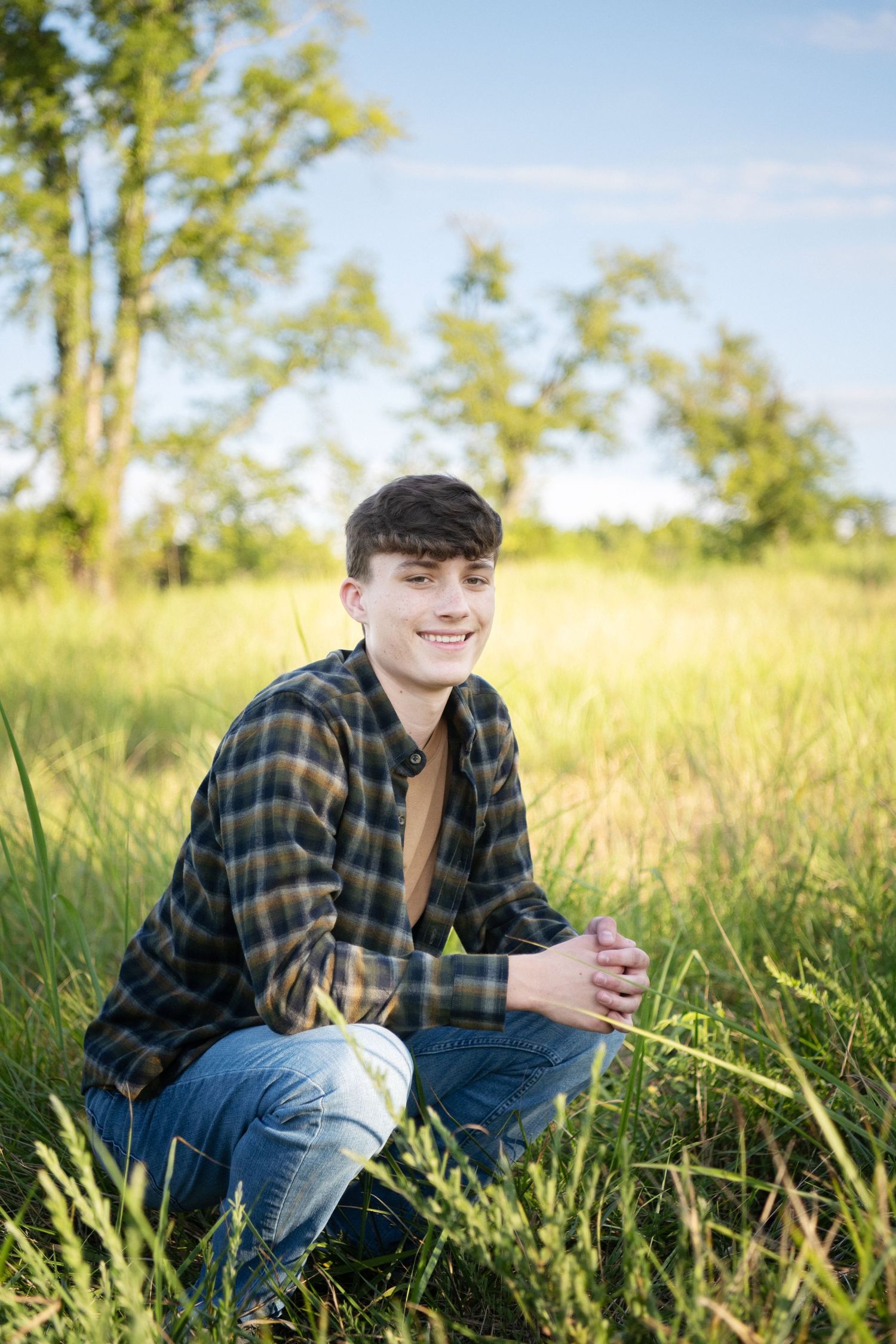A person wearing a plaid flannel shirt and jeans kneels in tall grass on a sunny day with trees in the background.