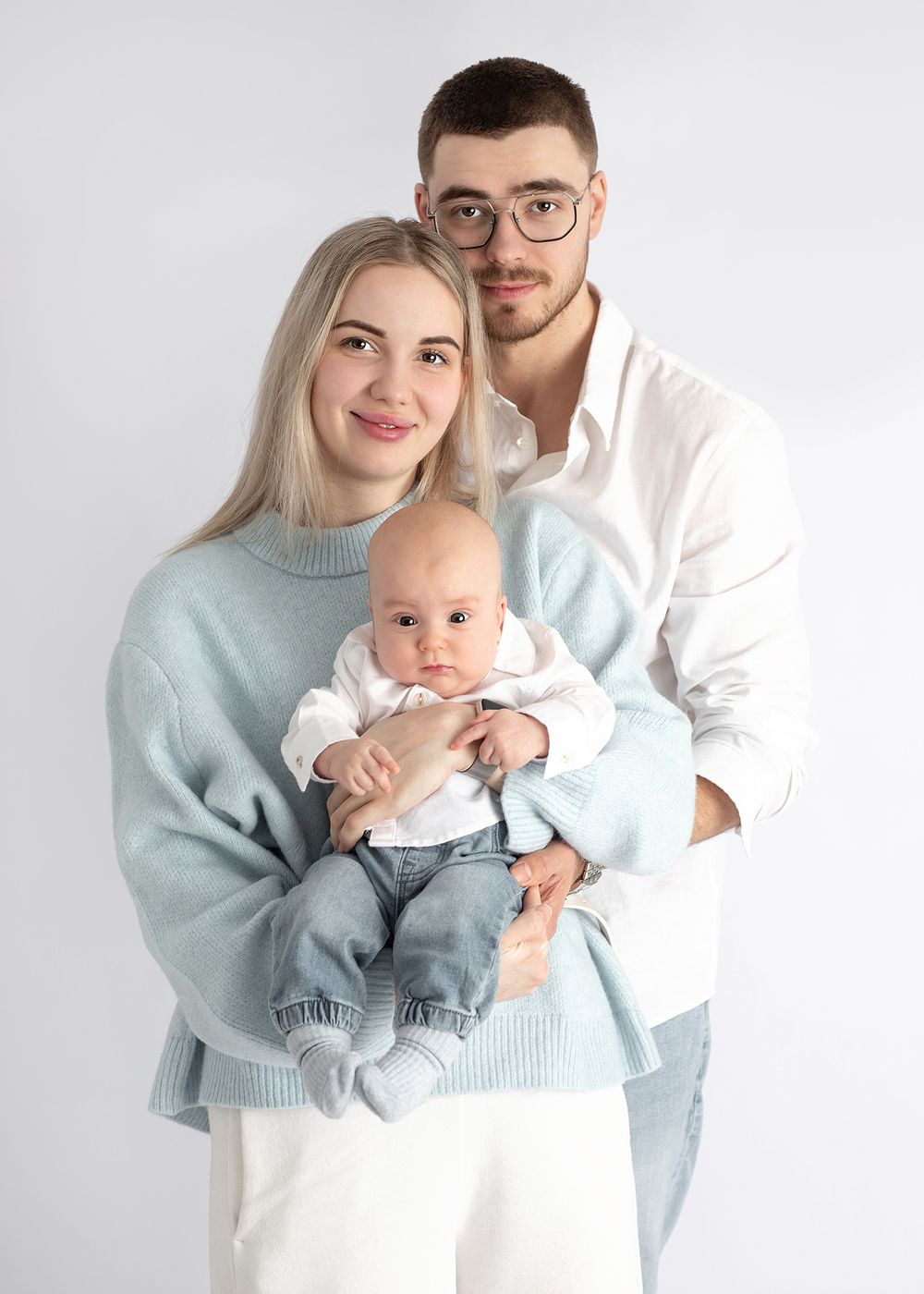 A couple poses with their baby wearing casual outfits against a white background in a studio setting.