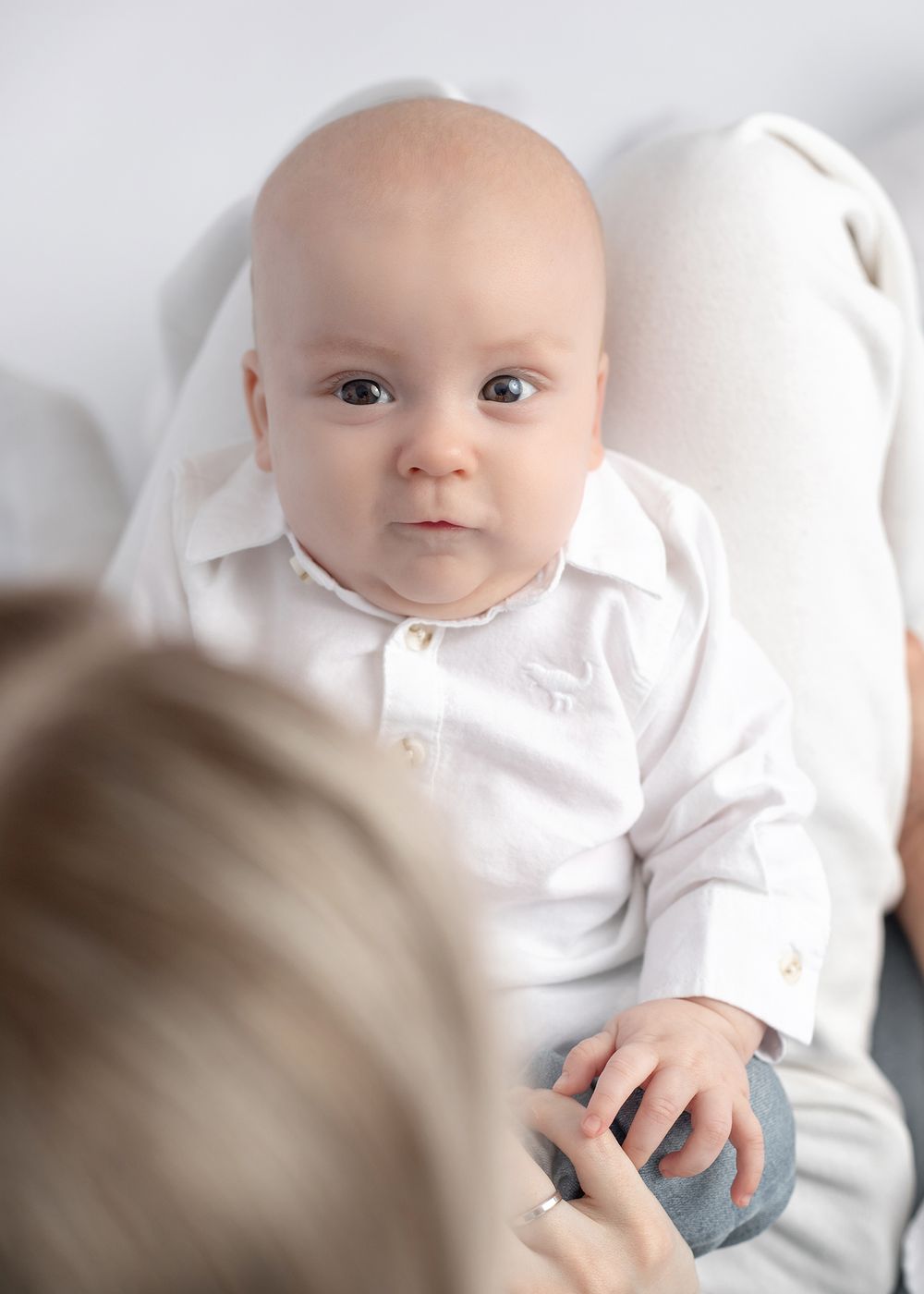 Baby wearing white long sleeve shirt sits against light background looking at camera.