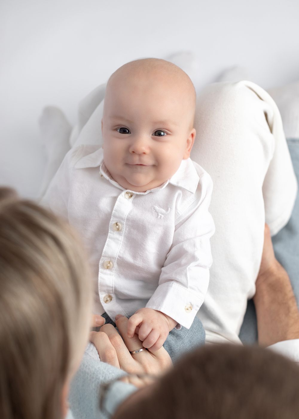 A baby in a white button-up shirt sits on someone's lap against a light background.