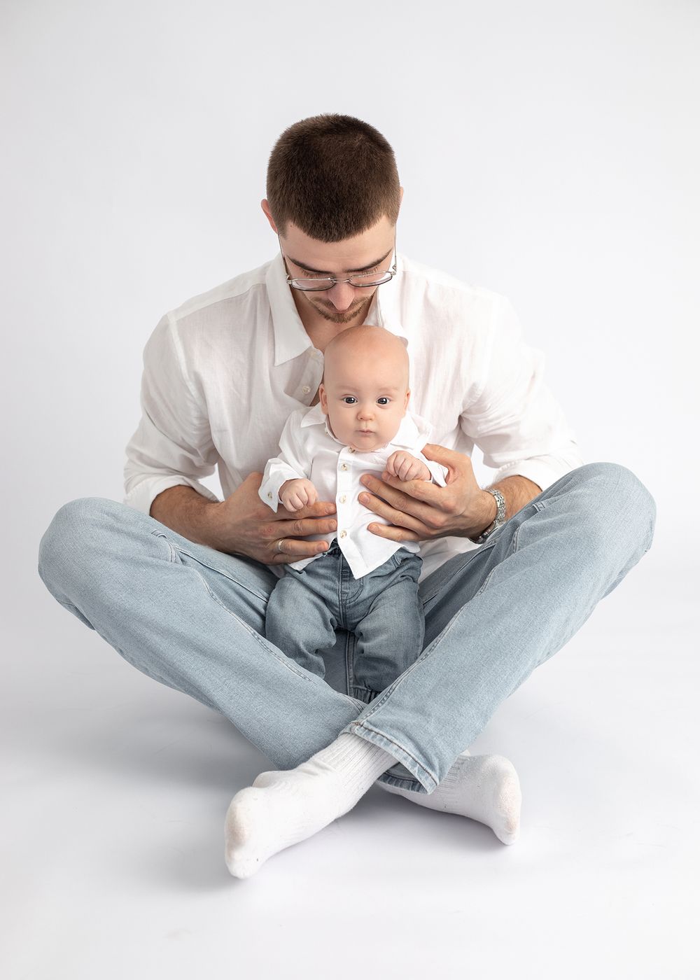 Parent in white shirt and jeans sits cross-legged holding infant in matching white outfit.