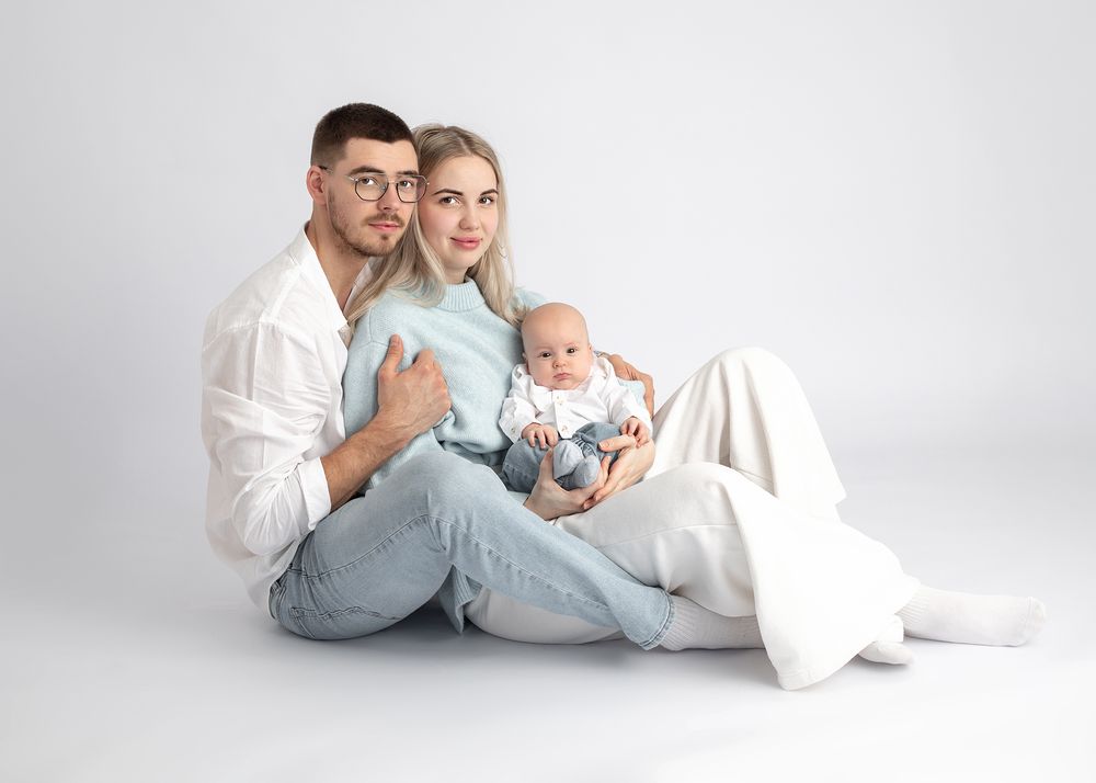 Family sitting together on white floor with baby wearing light blue outfit in studio setting.