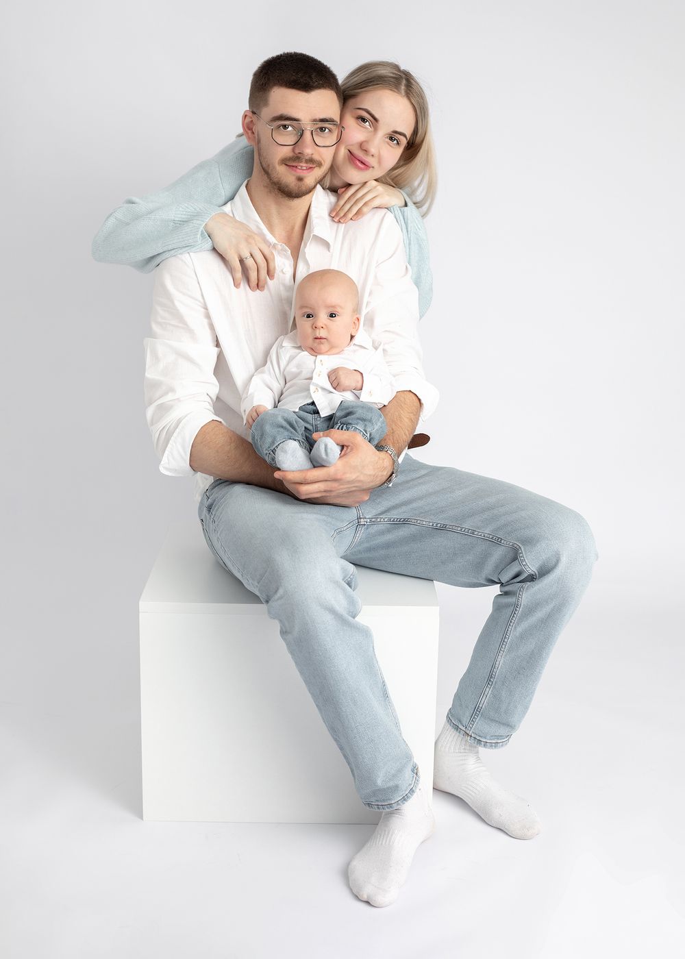 A couple in light-colored clothing sits together with their baby against a white backdrop in a series of casual poses.