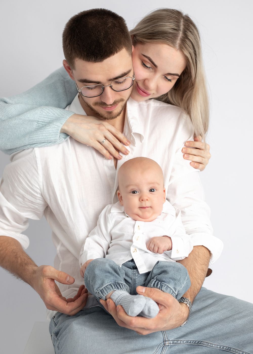 Parents in white and blue outfits cuddle with their baby on a light background in a series of tender moments.