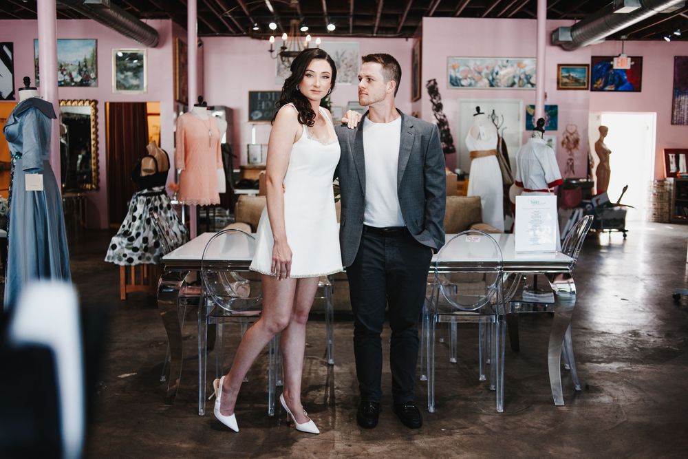 Couple posing in a boutique dress shop, woman in white mini dress and heels, man in gray blazer and black pants.
