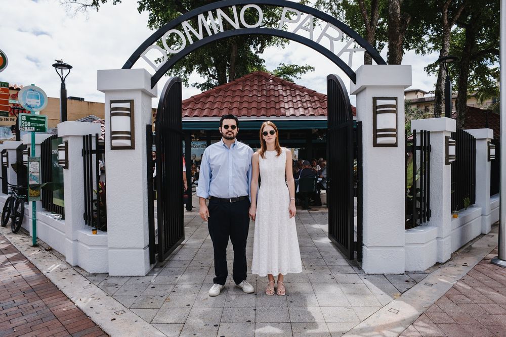 Couple in formal attire standing before an ornate gate entrance at Domino Park in Miami.
