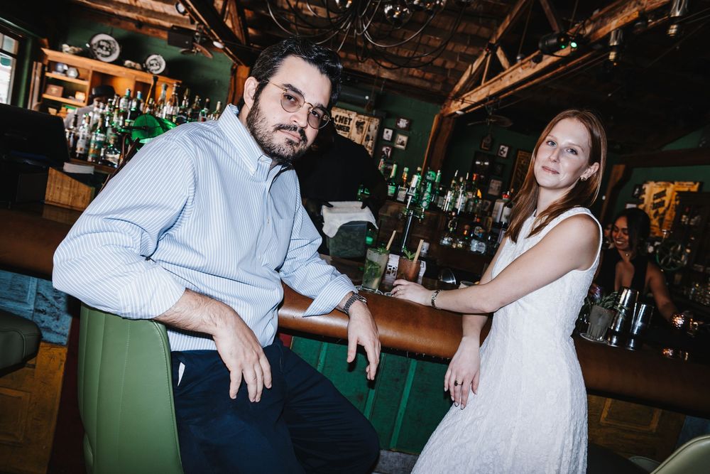A man and woman pose casually at a stylish green bar with bottles in the background.