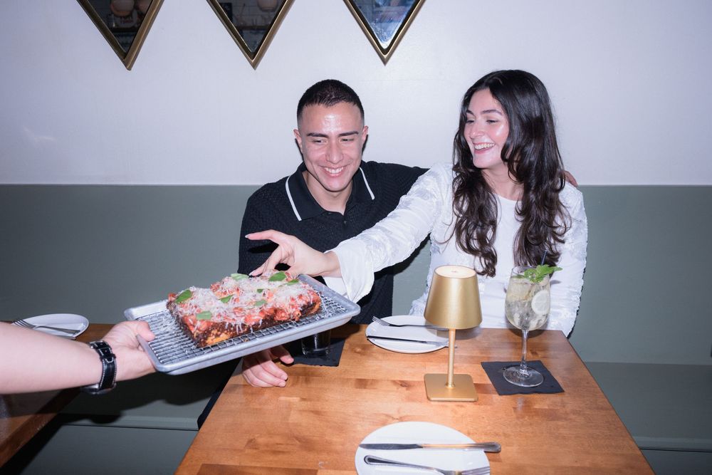 Couple smiling as a server presents a delicious dish at a restaurant table with cocktails.