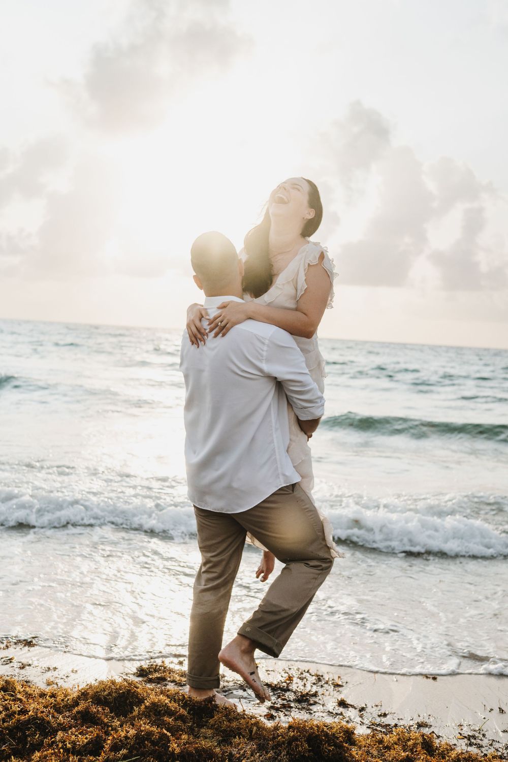 Couple embracing on a beach at sunset, man lifting woman in white dress near ocean waves.