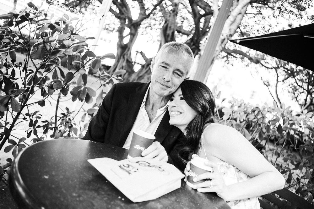Black and white photo of a couple sitting together at an outdoor table, sharing a tender moment in a garden setting.