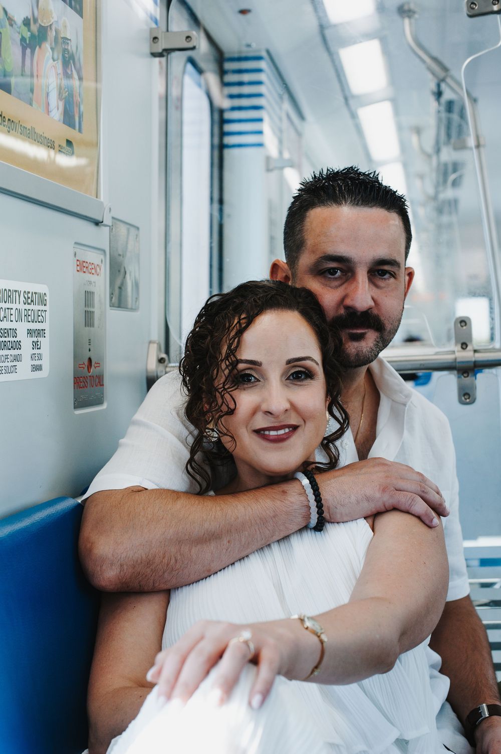 A couple dressed in white embraces while sitting on blue seats in what appears to be a subway car or train.