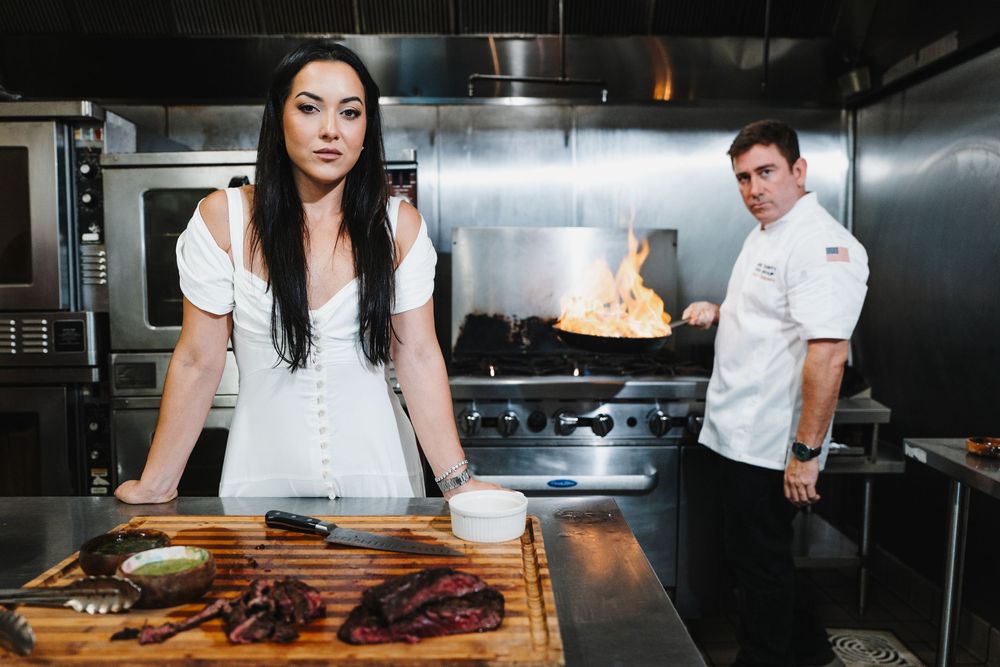 Chef in white uniform prepares food at stainless steel kitchen counter while other staff members work in background.