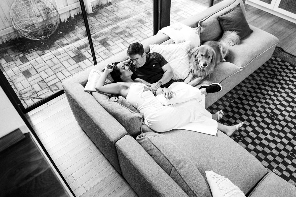 Black and white photo showing a person relaxing on a couch with a dog while reading a book.