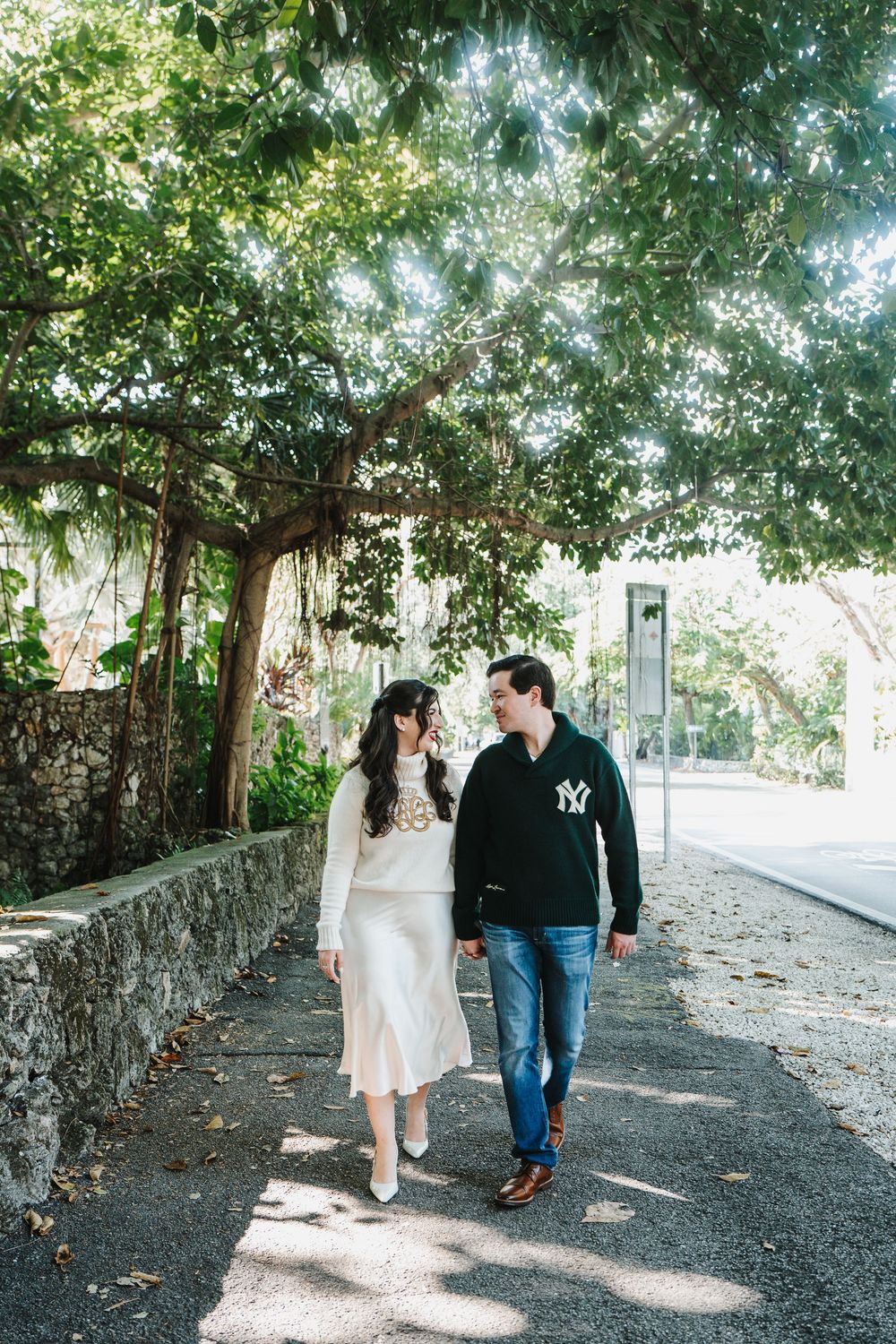 A couple walks together on a tree-lined path with dappled sunlight filtering through the leaves above.