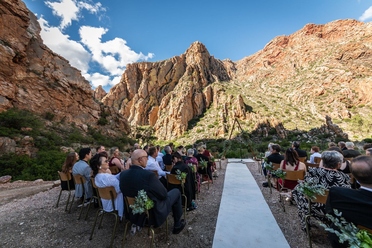Outdoor wedding ceremony in a stunning red rock canyon with guests seated along a white aisle under blue skies.
