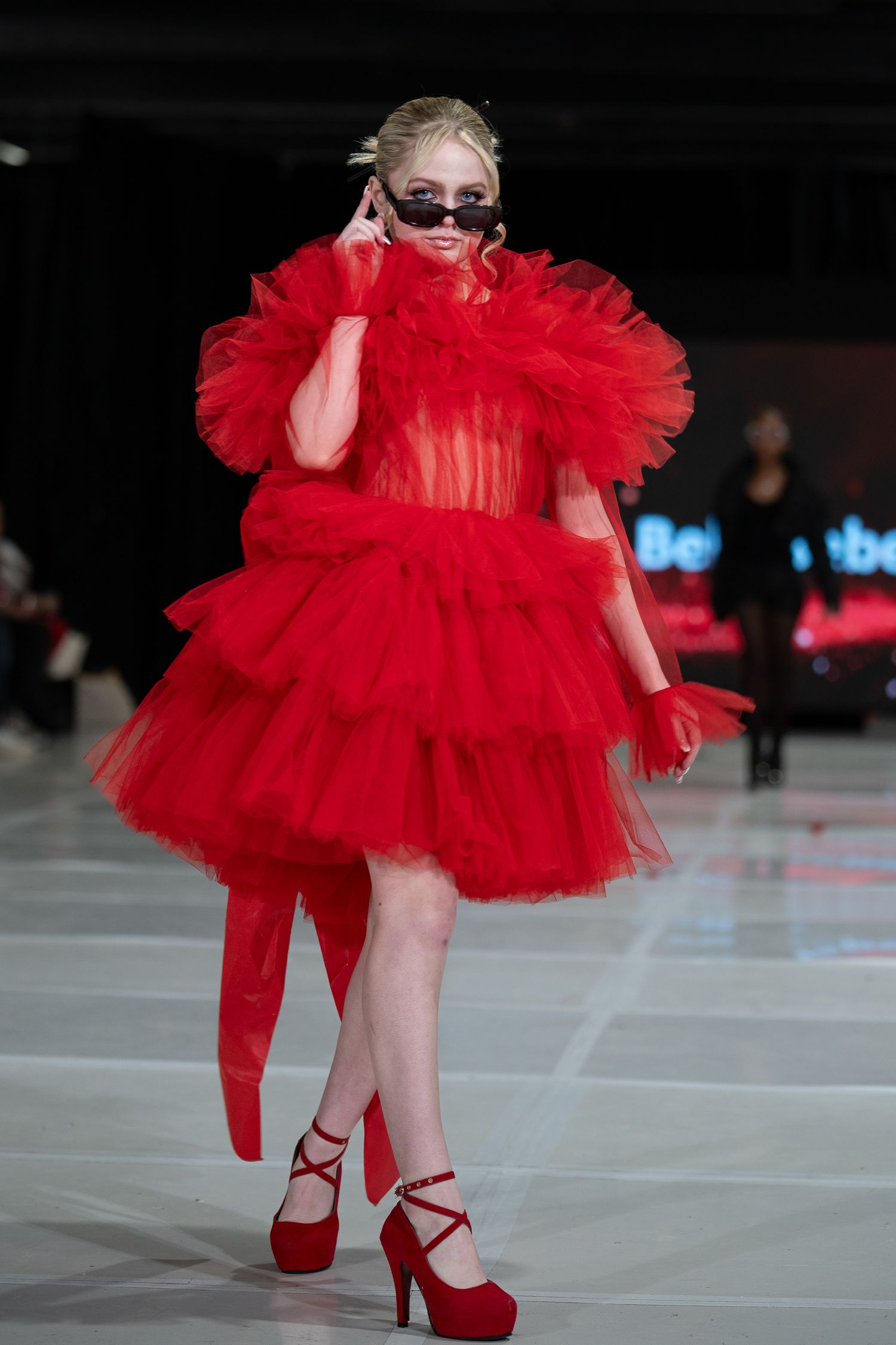 A model walks the runway in a dramatic red tulle mini dress with matching red platform heels during a fashion show.
