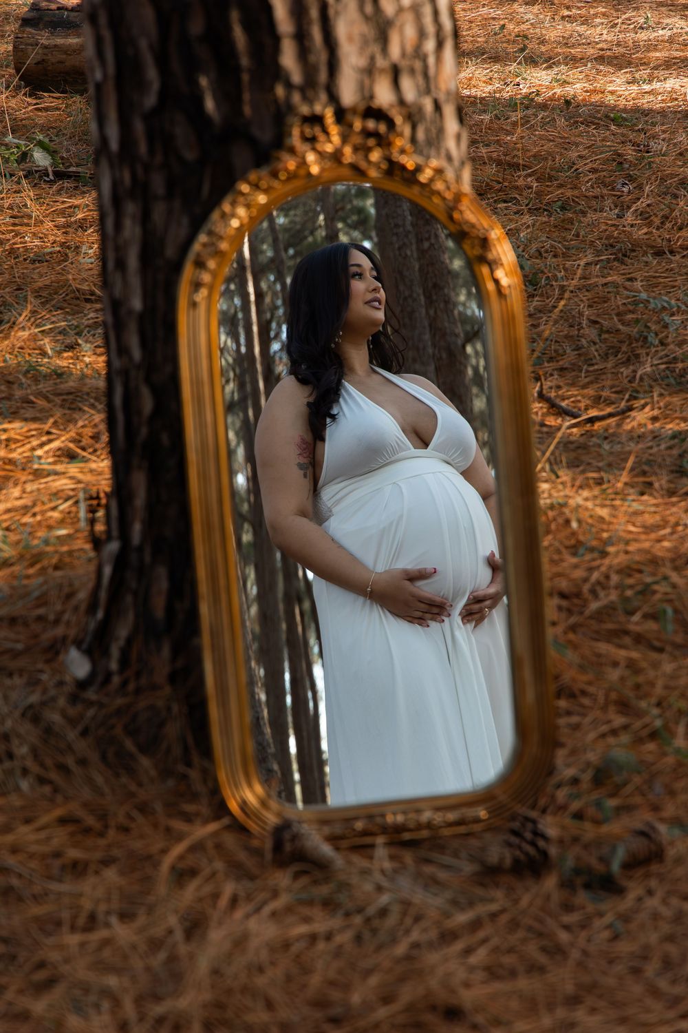 Artistic maternity portraits reflected in an ornate gold mirror placed among pine trees in a forest.