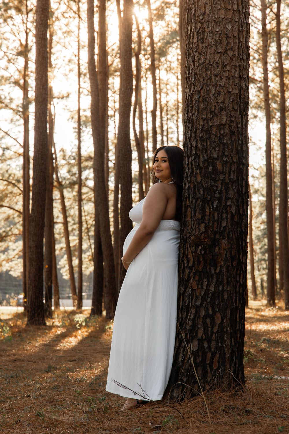 A figure in a flowing white dress leans against tall pine trees at sunset creating a dreamy forest scene.
