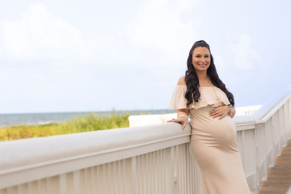A pregnant woman in a beige dress poses on a beach boardwalk during a maternity photoshoot.