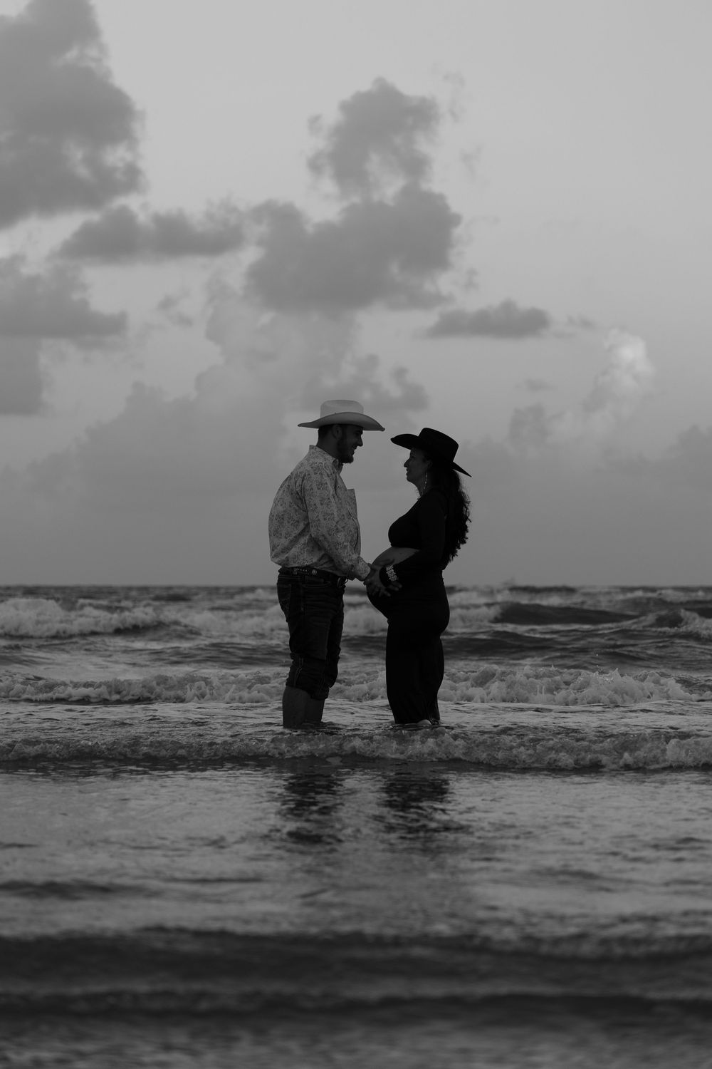 Two figures in traditional conical hats stand in ocean waves at dusk, creating a moody black and white silhouette.