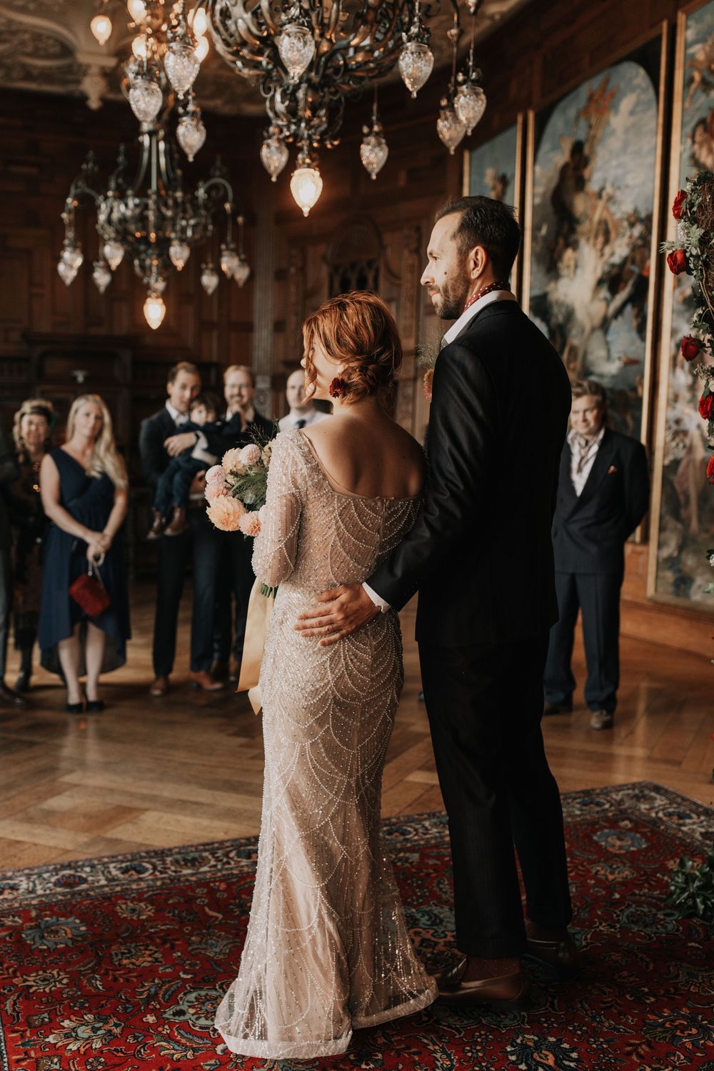 Elegant wedding ceremony in ornate room with vintage chandeliers as couple shares intimate moment on Persian rug.