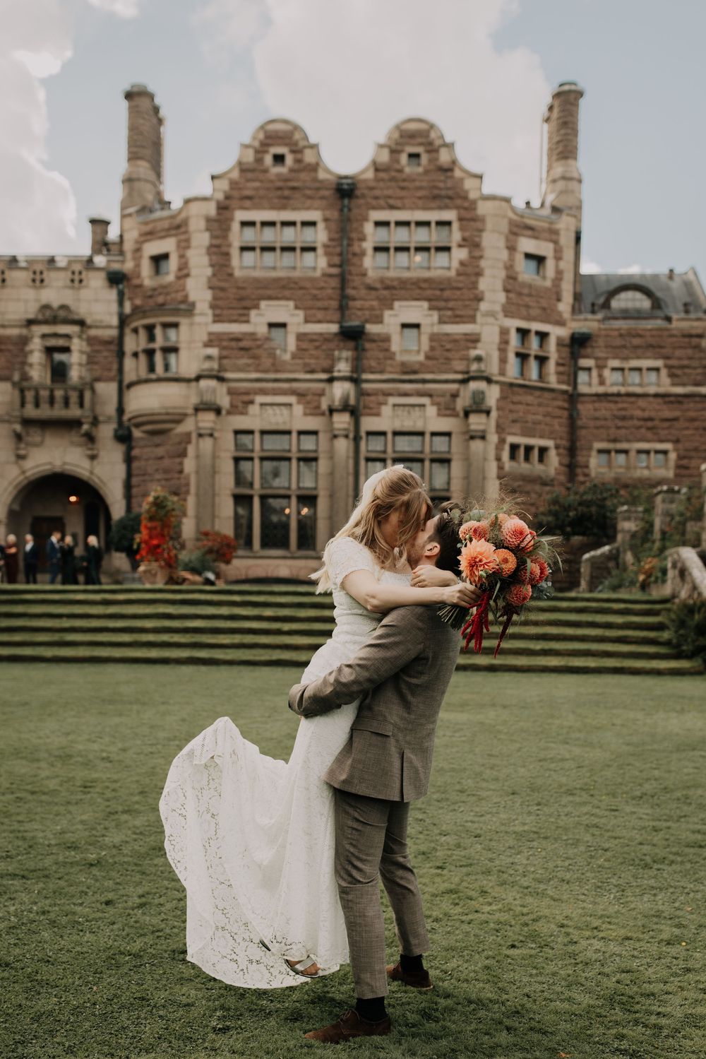 A couple dances and celebrates on a lawn in front of a grand Tudor-style mansion.