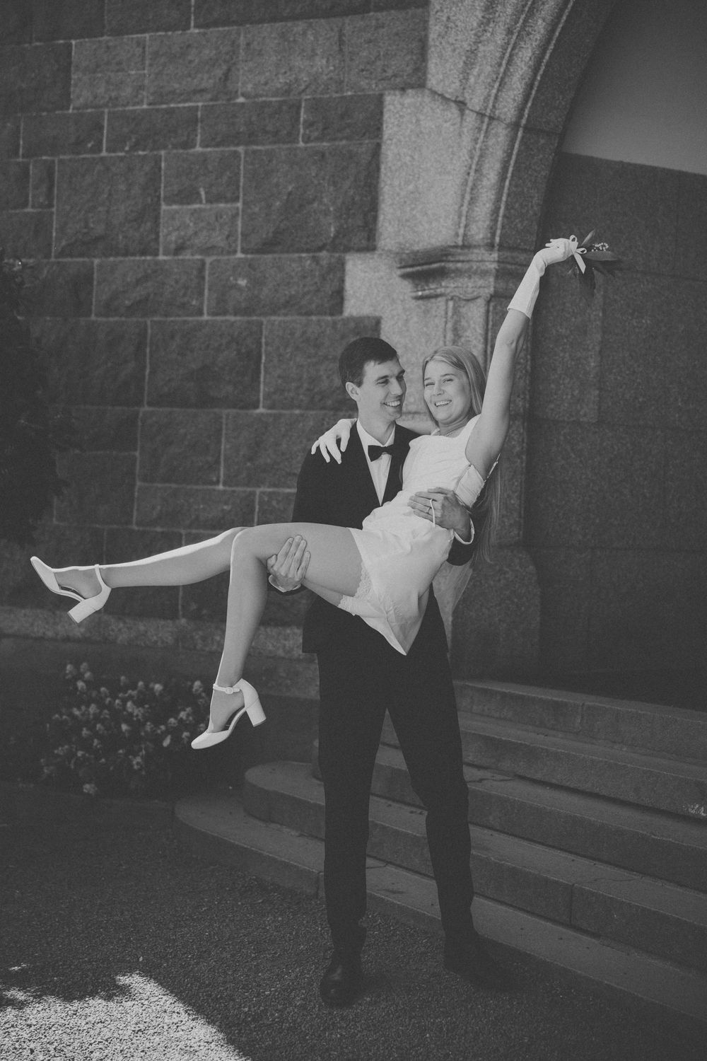 Groom in tuxedo carrying bride in white dress outside stone church in black and white wedding photo.