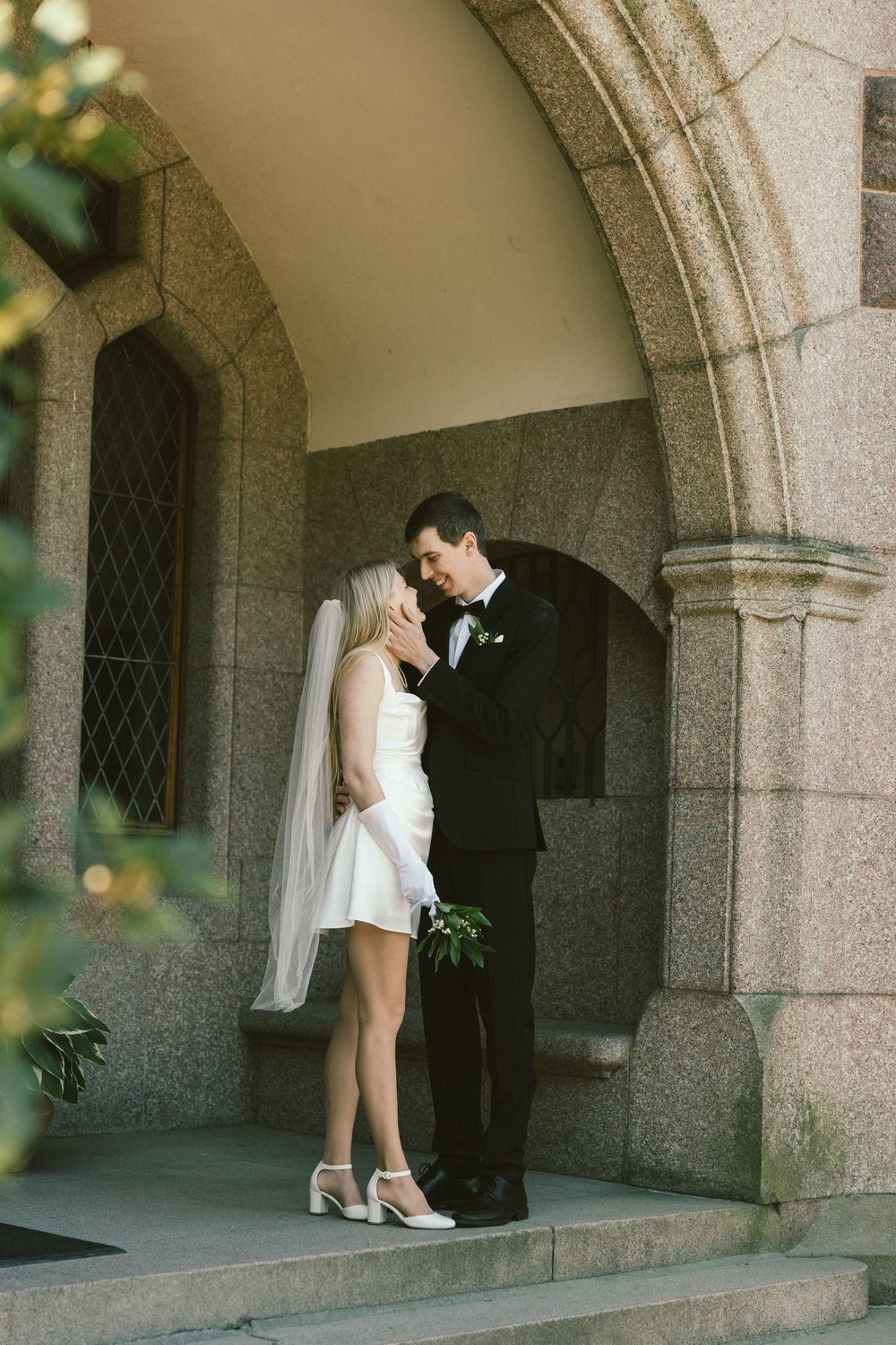 Bride in short white dress and veil kisses groom in black tuxedo on church steps under stone arch.