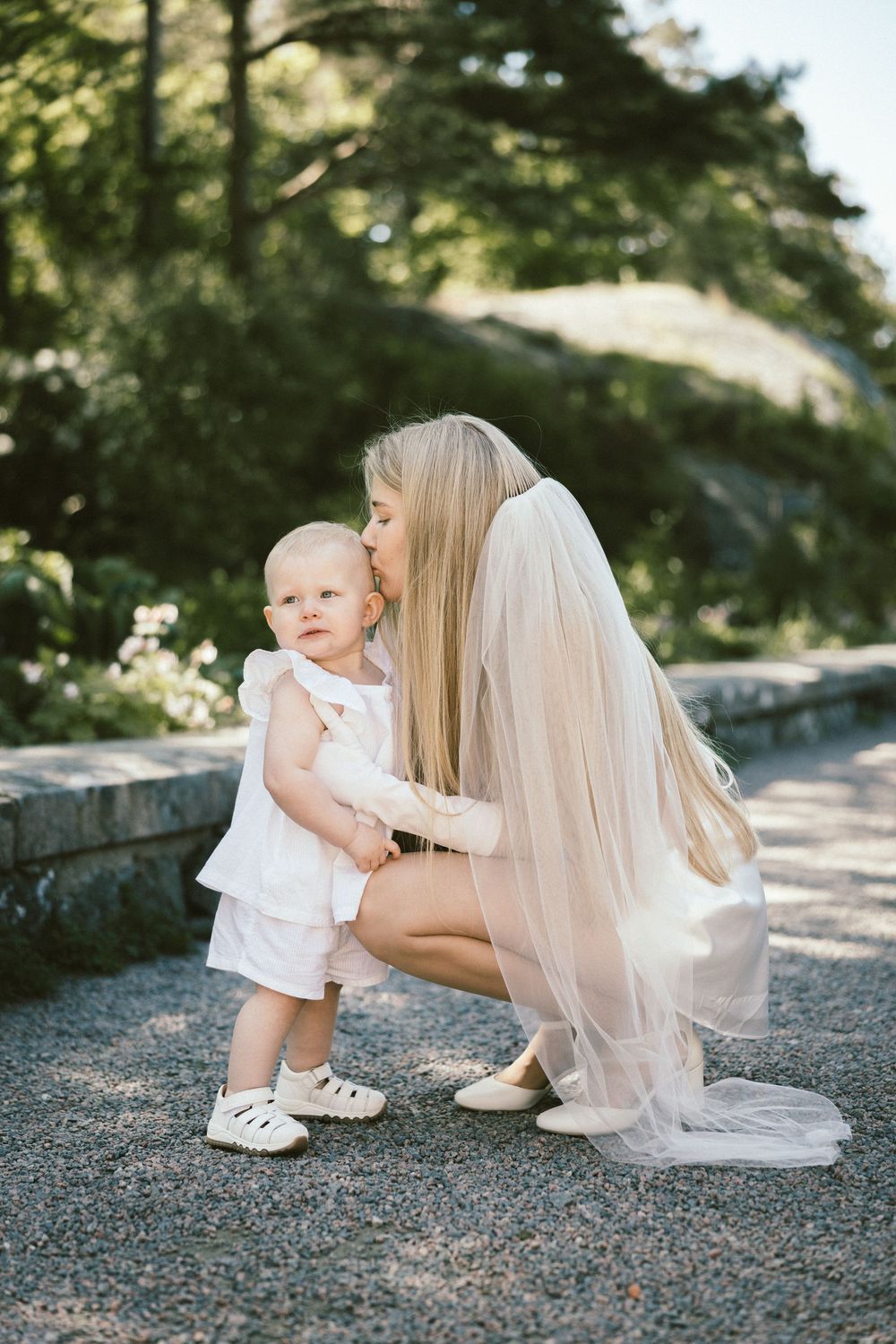 A bride in a white gown and veil crouches to kiss a baby in white on a sunlit garden path.
