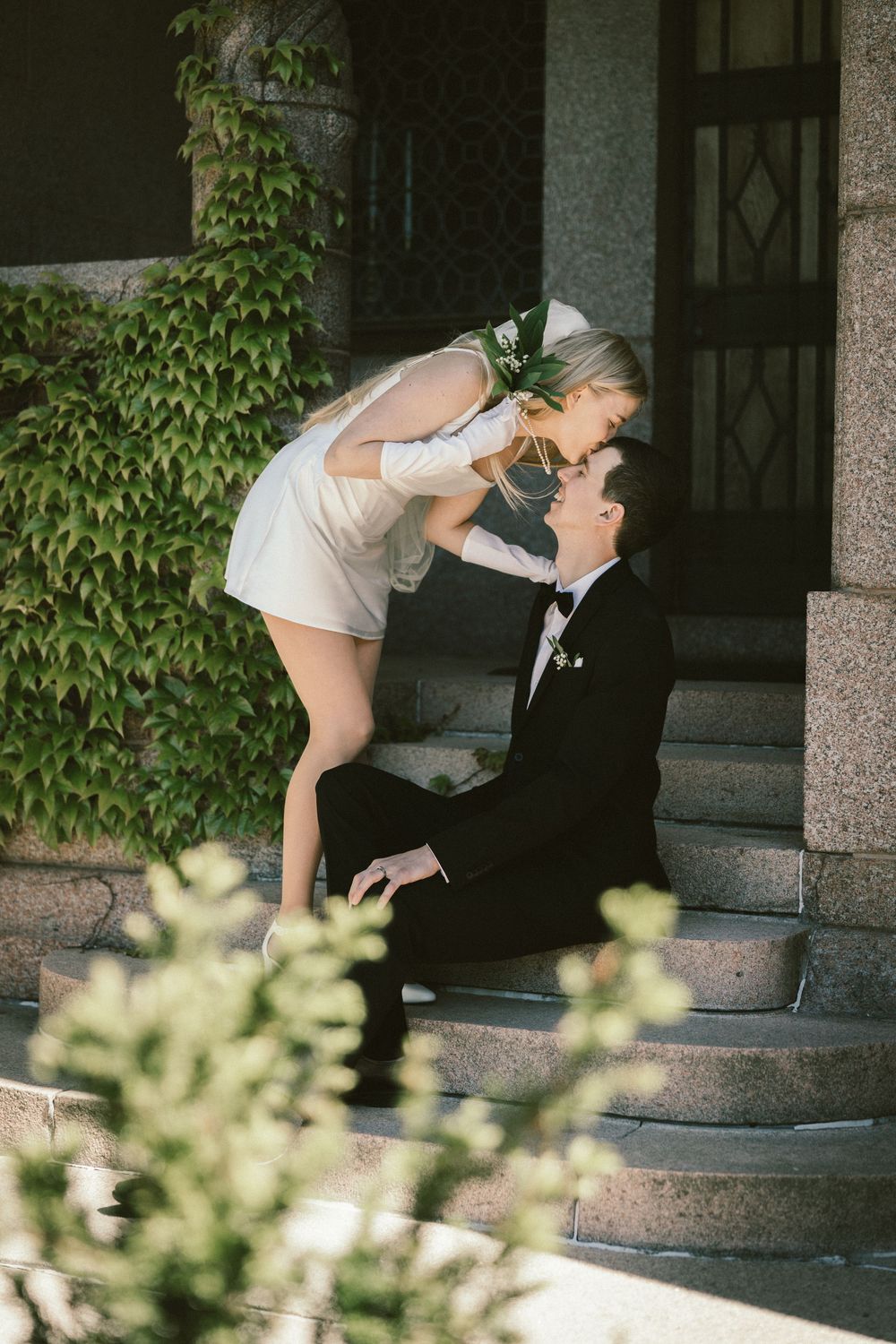 Bride in white mini dress leans down to kiss groom in black tuxedo sitting on stone steps with ivy backdrop.