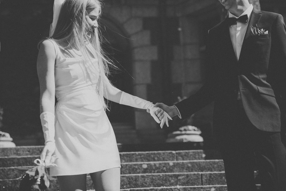 Black and white photo of a bride in a short white dress and groom in a tuxedo holding hands on stone steps.