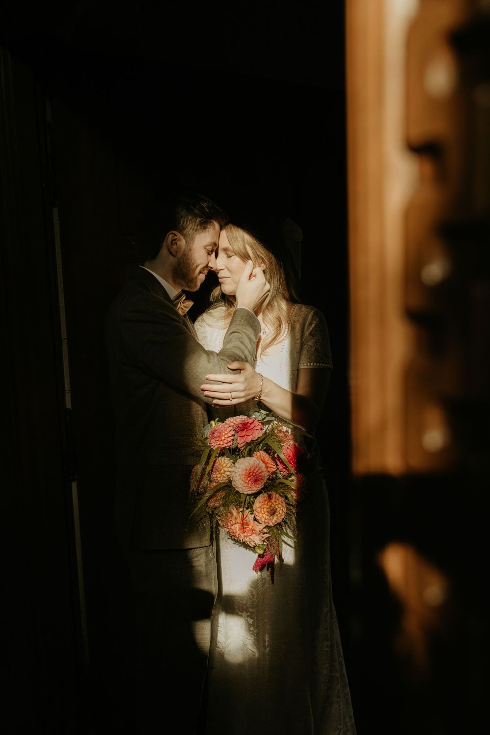 A romantic wedding couple shares an intimate moment in dramatic lighting while holding a coral floral bouquet.