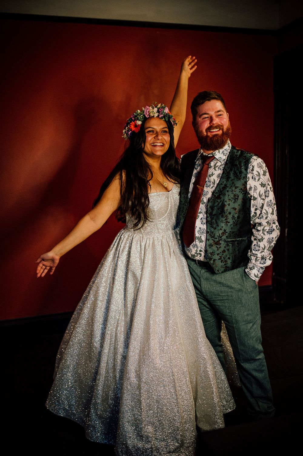 A couple poses for formal portraits against a dark red backdrop wearing a sparkly white dress and green vest.