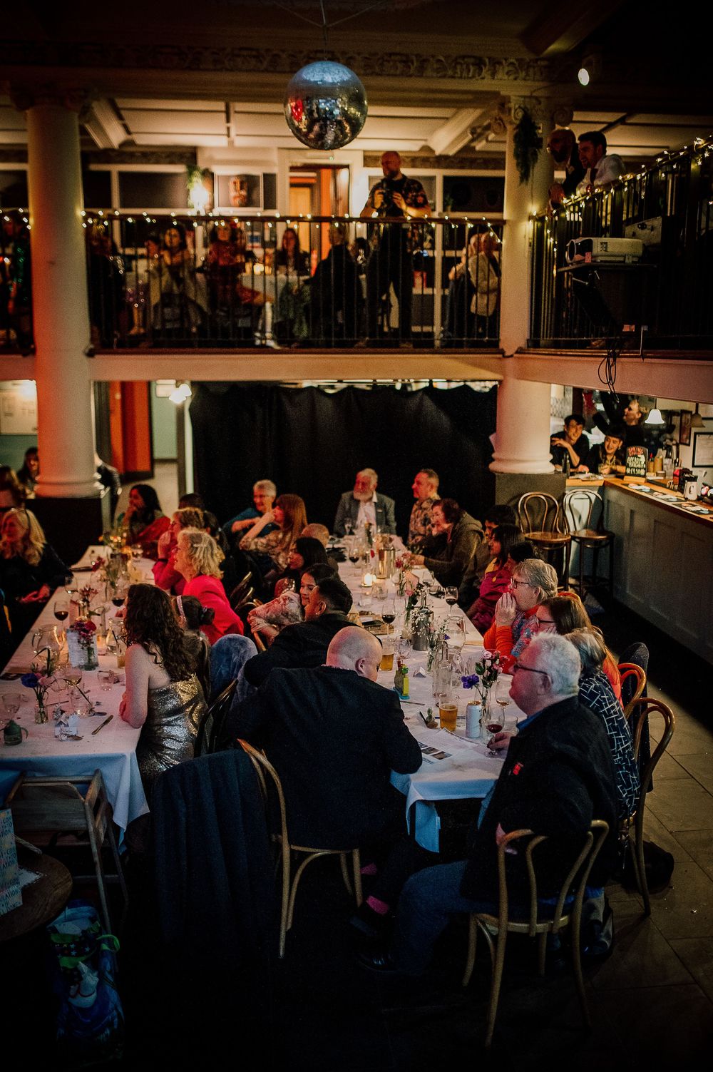 Wedding reception banquet hall with multiple round tables of guests dining under ambient lighting.