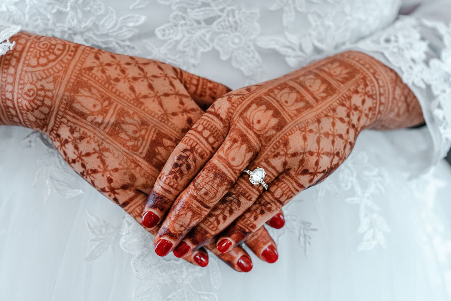 Bride's hands adorned with intricate henna mehndi design and diamond ring, red nail polish, white lace dress.