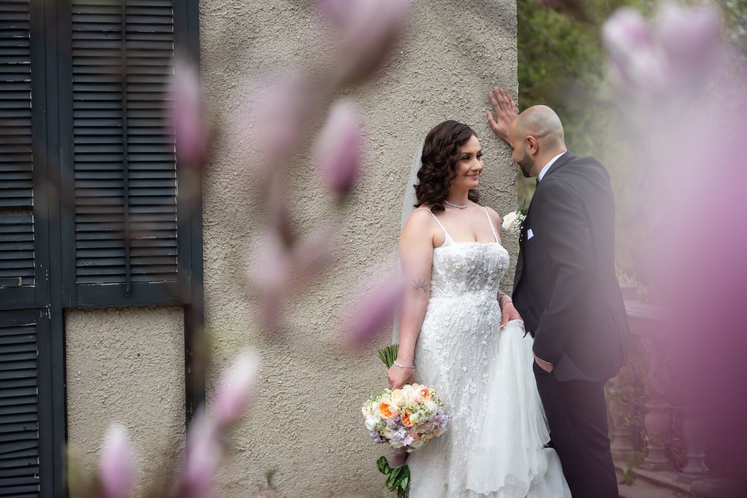 Bride in lace gown holds colorful bouquet while groom look at her near blooming magnolia trees outdoors at Glendon College Gardens.