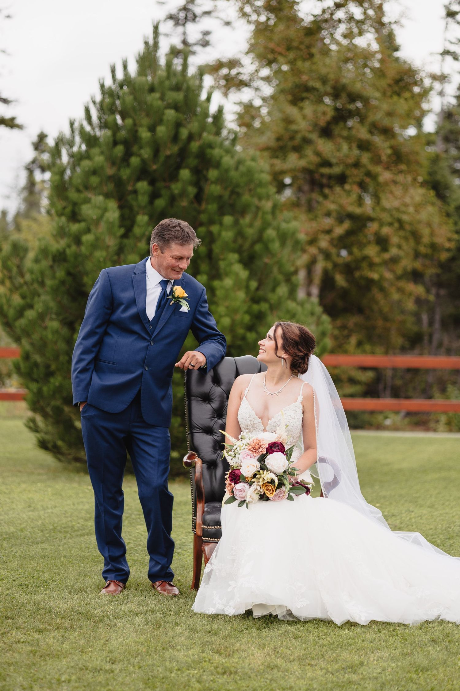 A couple in wedding attire sharing a tender moment on grass surrounded by pine trees.