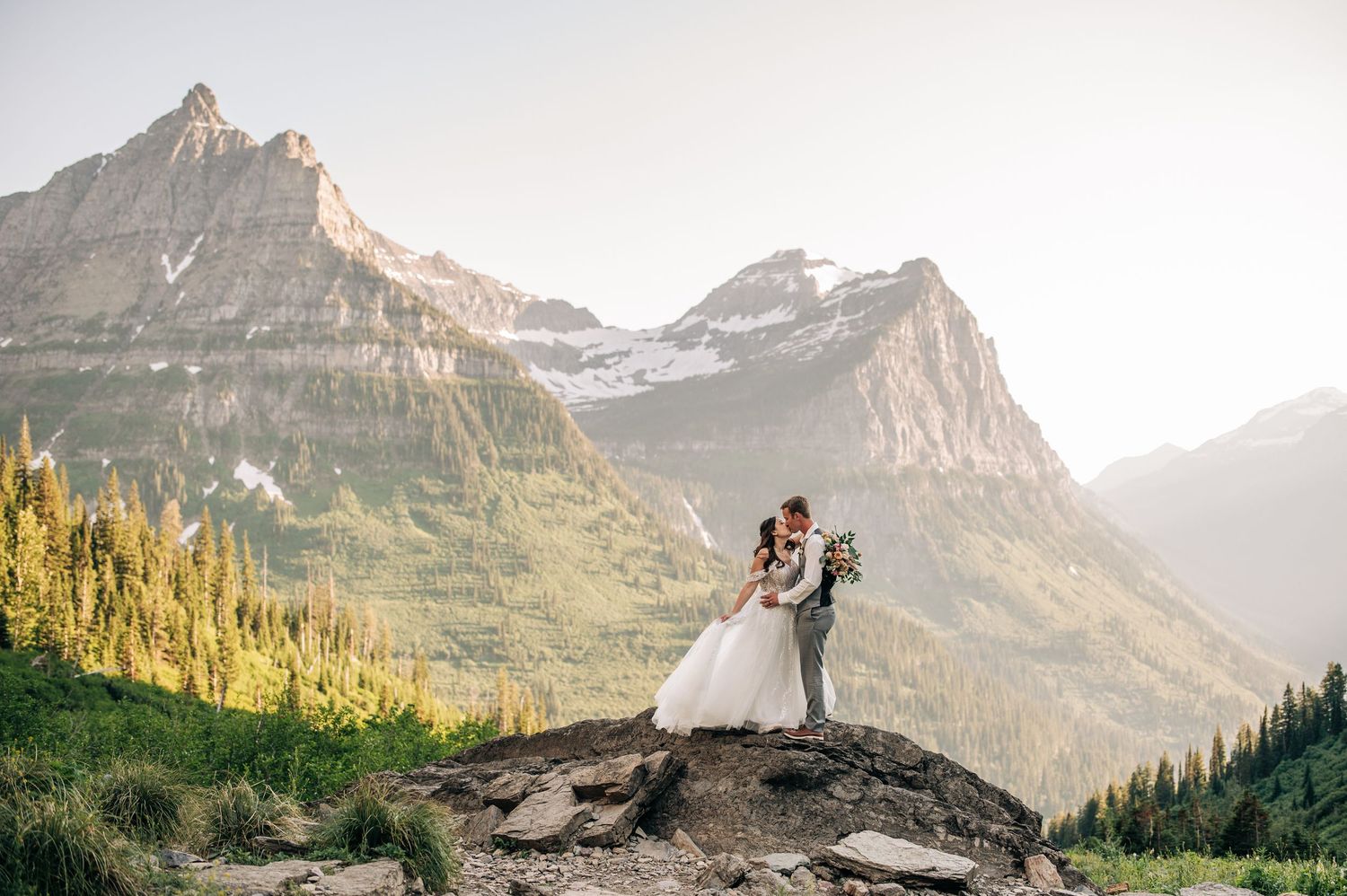Two brides hold hands by a mountain river surrounded by evergreen trees and misty peaks in Glacier National Park.