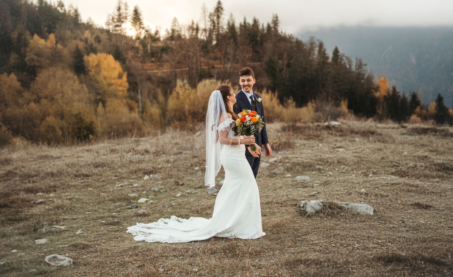 Happy couple in wedding attire smiling outdoors, bride holding white floral bouquet surrounded by lush greenery.