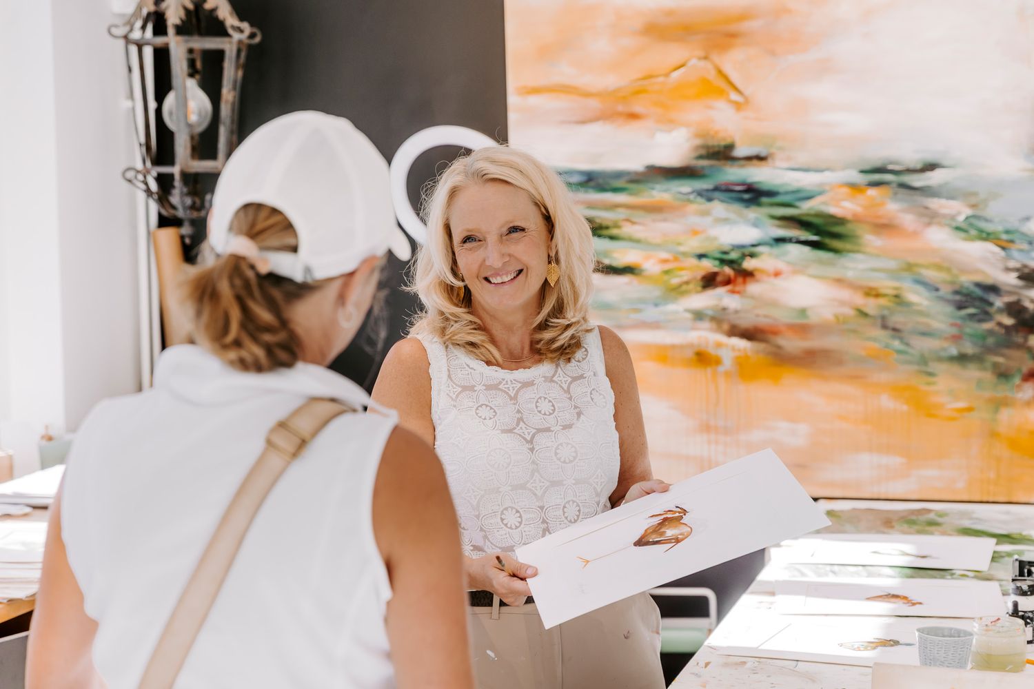 To kvinder i hvide outfits udveksler dokumenter i et lyst kunstgalleri med et farverigt abstrakt maleri. Fotograf Middelfart