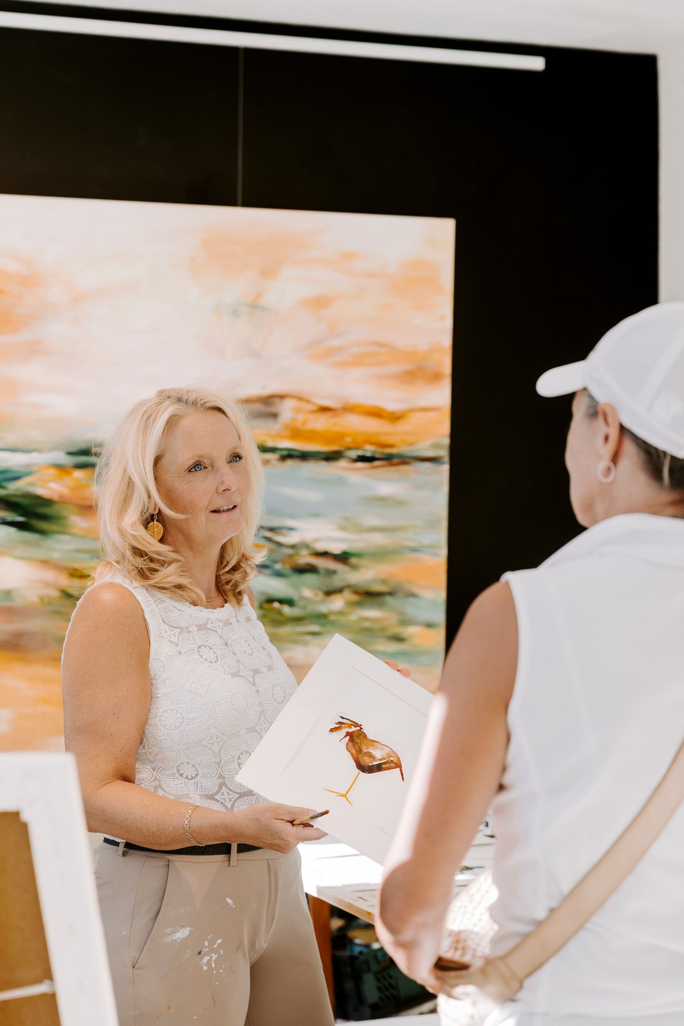 To kvinder i hvide outfits udveksler dokumenter i et lyst kunstgalleri med et farverigt abstrakt maleri. Fotograf Middelfart