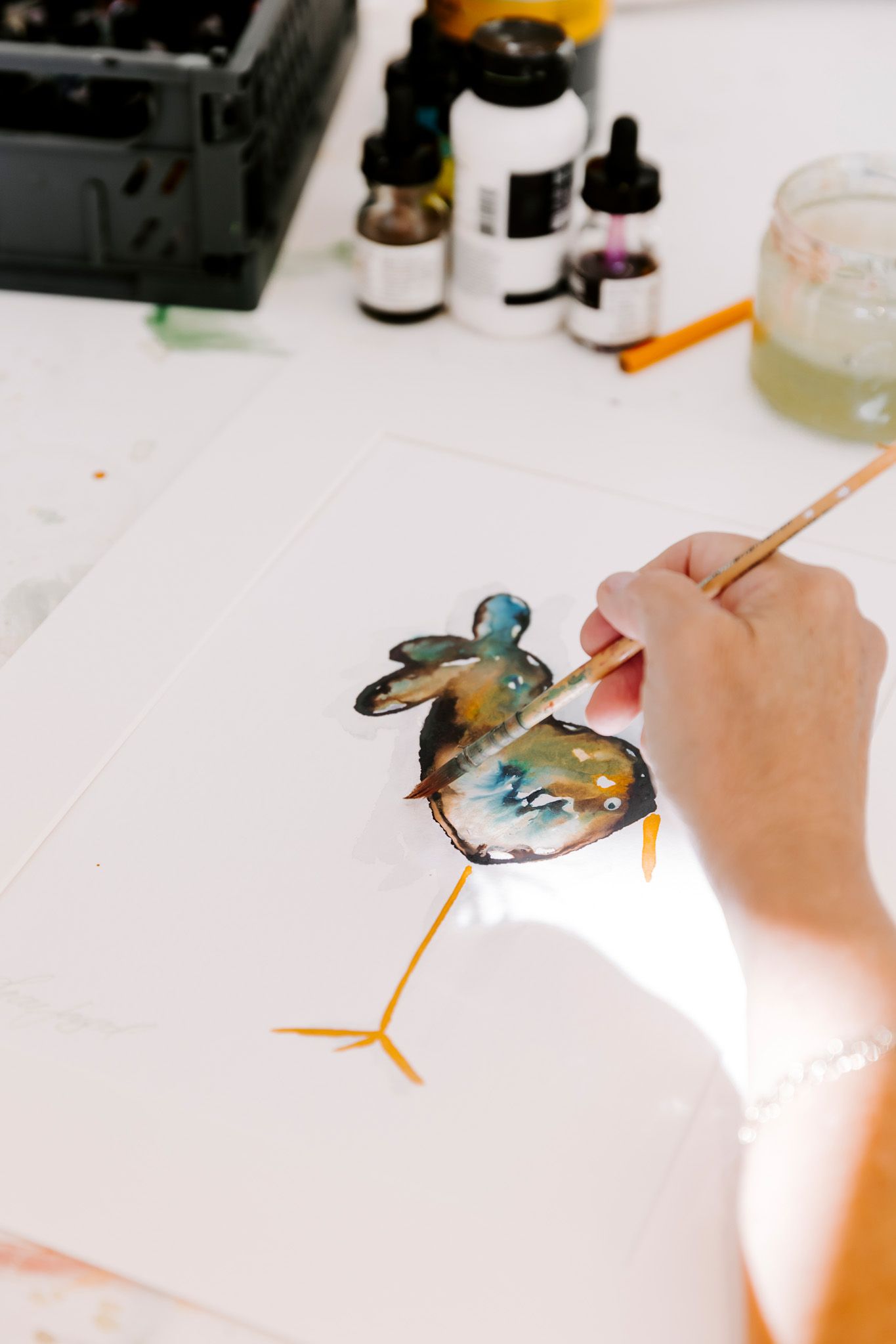 Kunstner maler en farverig fugleillustration på hvidt papir med akvarel og blæk ved et lyst studiebord. Fotograf Middelfart.