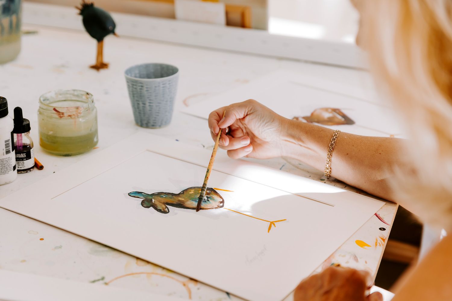 Kunstner maler en farverig fugleillustration på hvidt papir med akvarel og blæk ved et lyst studiebord. Fotograf Middelfart.