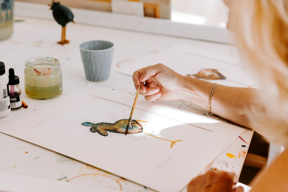 Kunstner maler en farverig fugleillustration på hvidt papir med akvarel og blæk ved et lyst studiebord. Fotograf Middelfart.