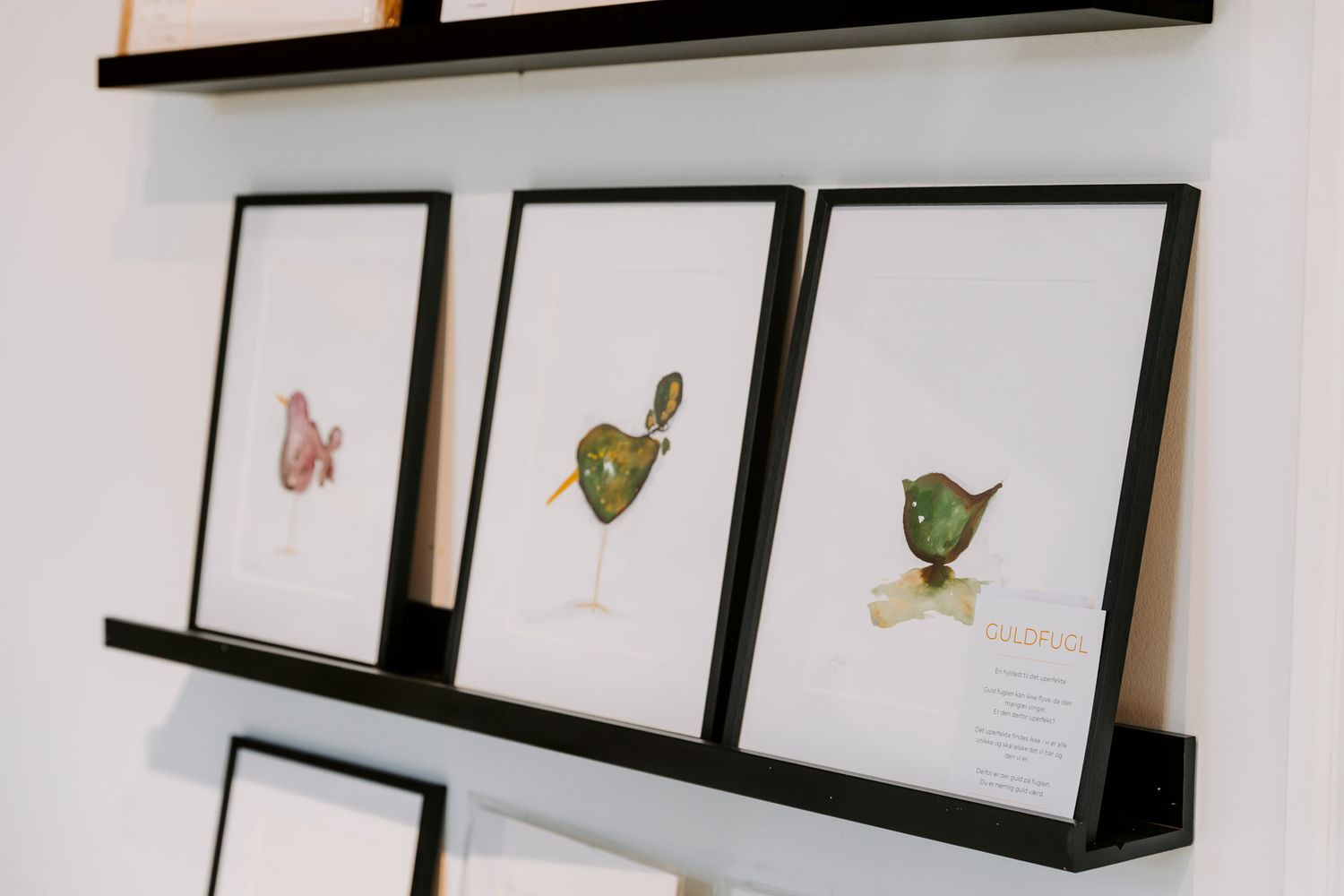 Tre indrammede botaniske cocktailillustrationer udstillet på sorte væghylder i et moderne gallerimiljø. Fotograf Middelfart
