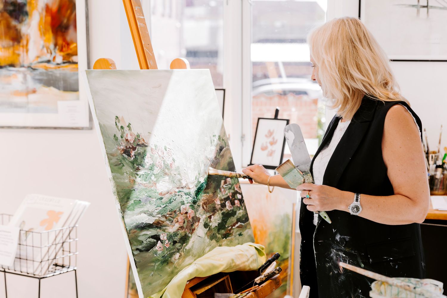Kunstner står i sit atelier ved siden af et staffeli og holder pensler, med store abstrakte orange malerier i baggrunden. FotografMiddelfart