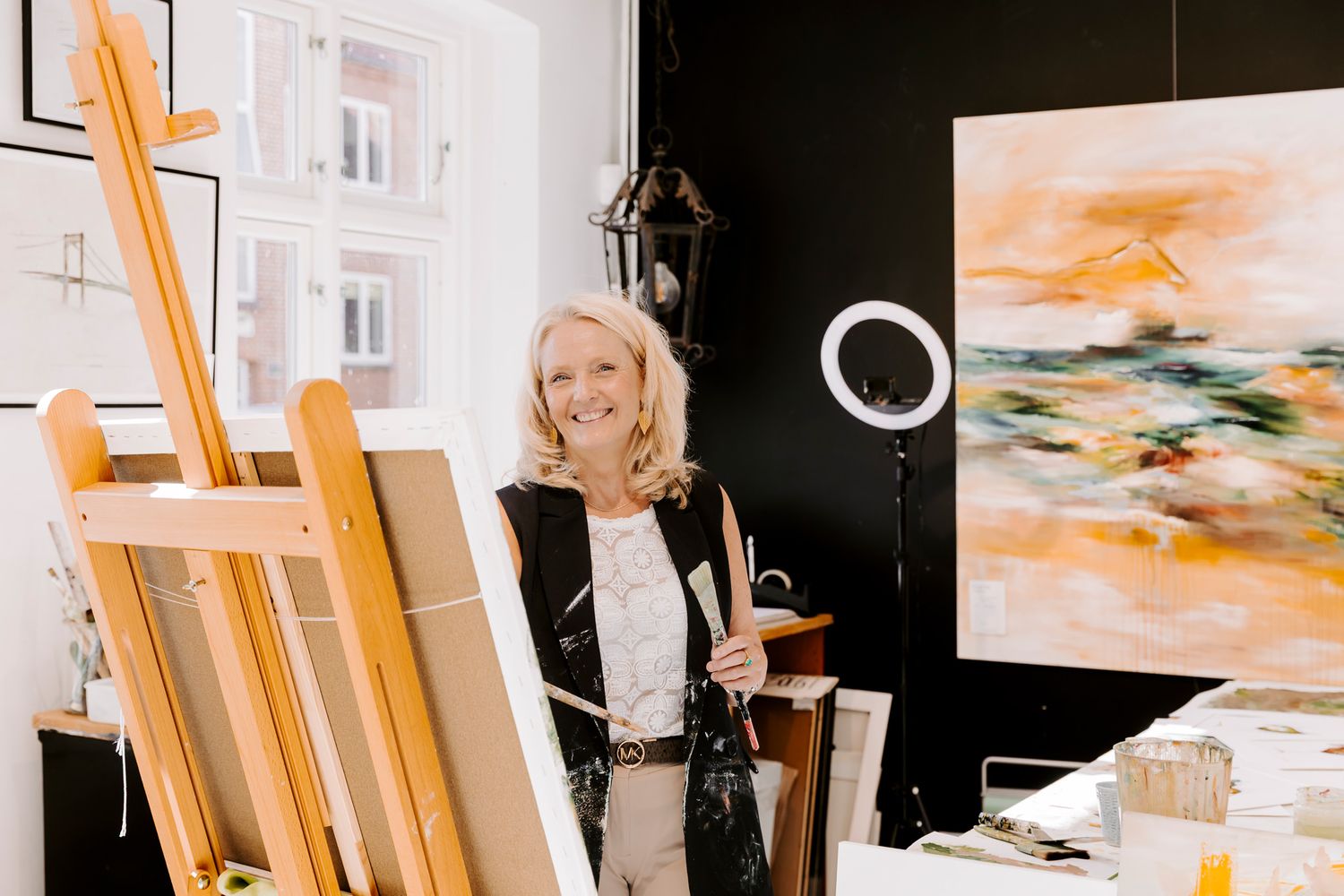 Kunstner står i sit atelier ved siden af et staffeli og holder pensler, med store abstrakte orange malerier i baggrunden. FotografMiddelfart