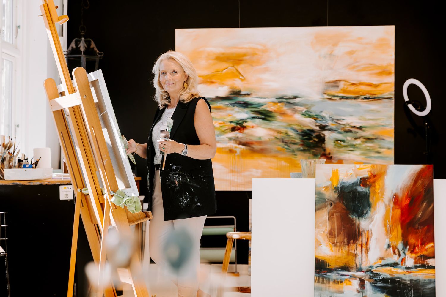 Kunstner står i sit atelier ved siden af et staffeli og holder pensler, med store abstrakte orange malerier i baggrunden. FotografMiddelfart