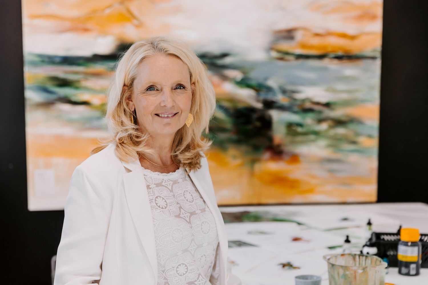 Kunstner står i sit atelier ved siden af et staffeli og holder pensler, med store abstrakte orange malerier i baggrunden. FotografMiddelfart